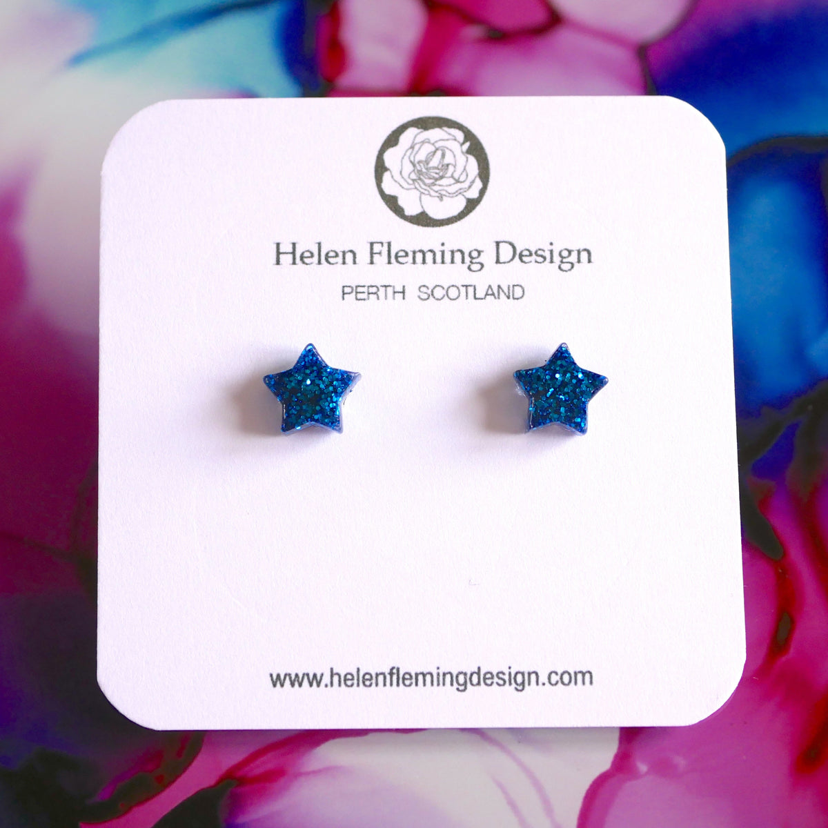 A pair of small blue glitter stud earrings displayed on a branded card, Helen Fleming Design, with a colorful background.